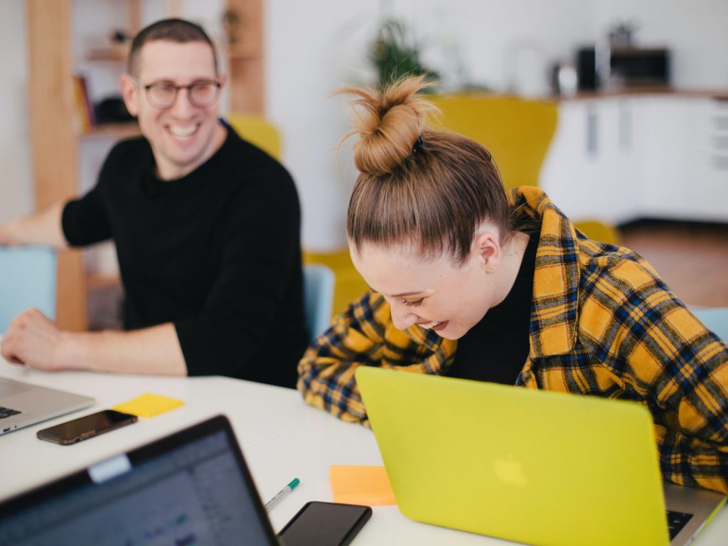 Photograph of a man and woman sat at a desk, woman laughing and using a yellow laptop