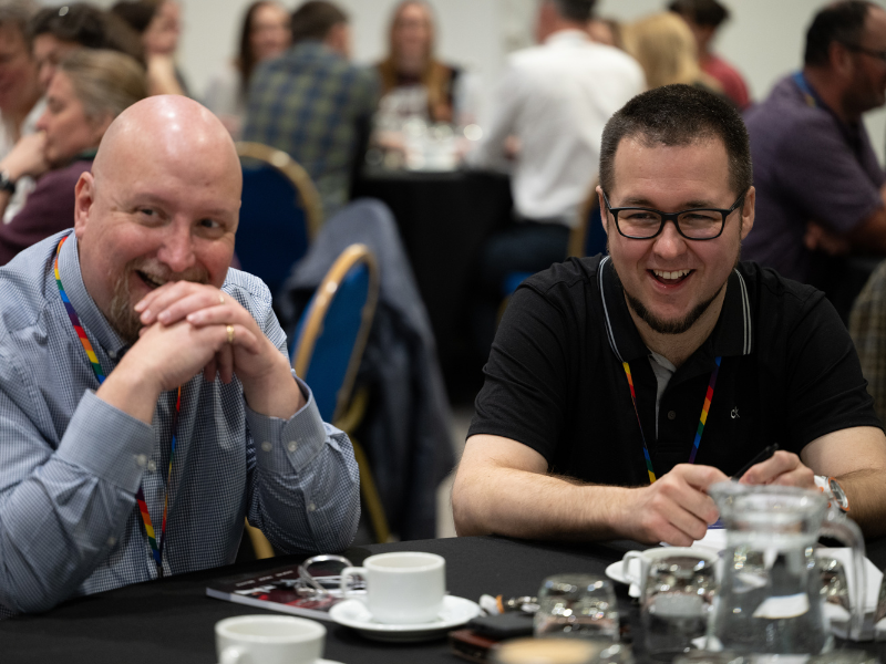 Photograph of two men at an event, sat at table, laughing.