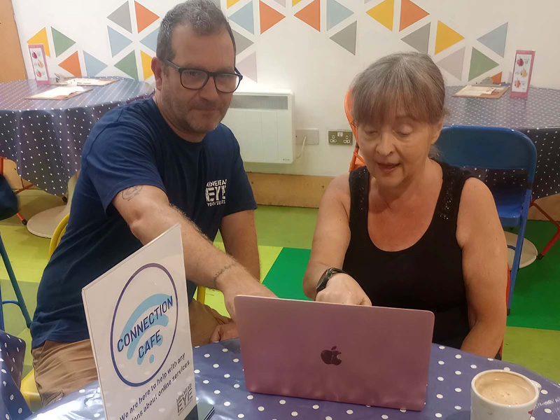 A man and a woman sitting at a table, looking at a laptop together in a community space.