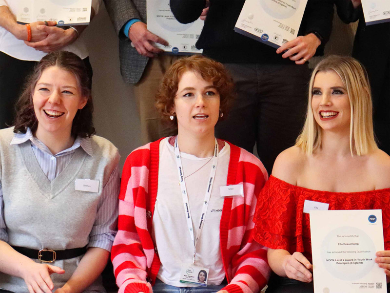 Three women sitting together, smiling and holding certificates during an awards ceremony.