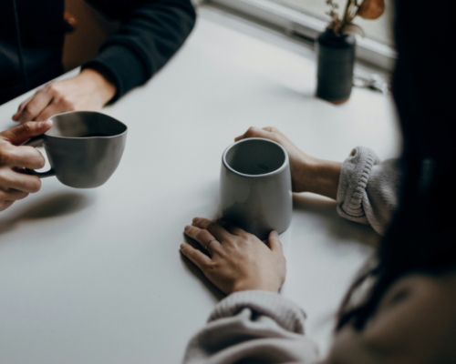 A close up of two people's hands as they sit at a table. They are holding mugs.