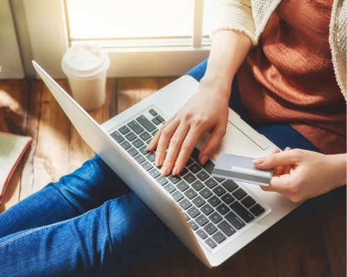 Photograph of a woman shopping online holding a credit card with a laptop on her lap, torso in shot.