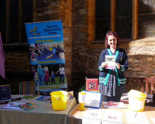 A member of the Taunton Opportunity Group standing at a table with donation buckets and leaflets. She is outside in the sunshine and smiling.